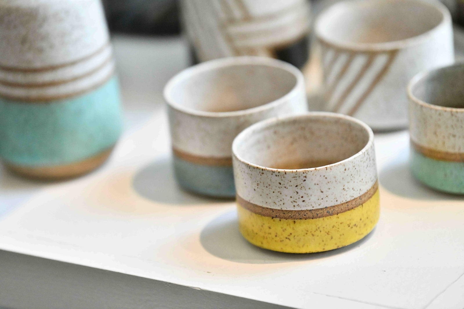 A close-up view of beautiful ceramic cups displayed at Frame Corner, a custom frame store, showcasing a variety of pastel-colored designs with smooth, minimalist patterns of blue, yellow, and white.